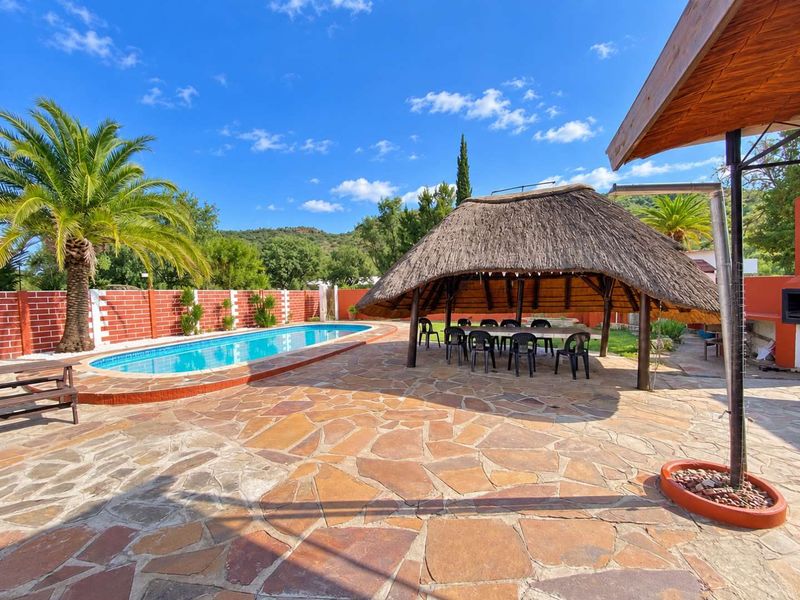Outdoor entertainment area featuring a swimming pool, a traditional thatched lapa, and stone paving, set against a scenic landscape.