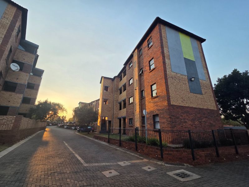 Exterior view of a multi-story face brick apartment complex featuring paved driveway access and perimeter security fencing at sunset.