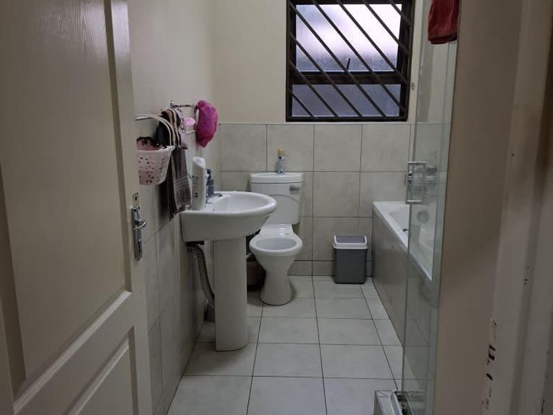 A functional bathroom featuring a pedestal basin, toilet, bathtub, and a glass-enclosed shower with neutral floor tiles and security bars on the window.
