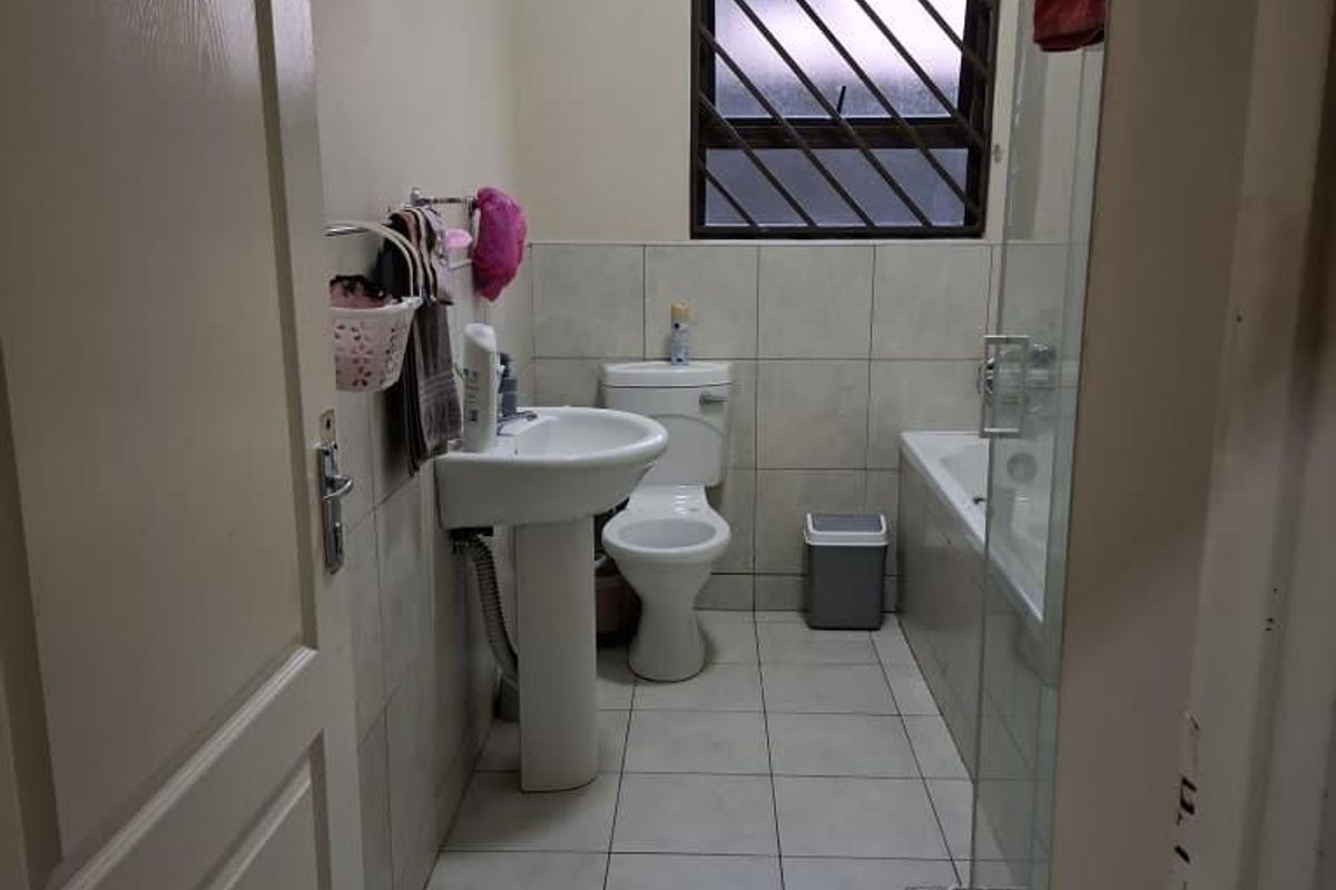A functional bathroom featuring a pedestal basin, toilet, bathtub, and a glass-enclosed shower with neutral floor tiles and security bars on the window.