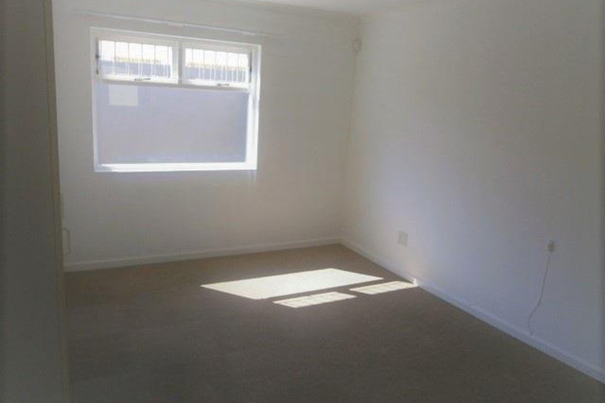A bright, empty bedroom featuring neutral carpeting and a window with security bars.