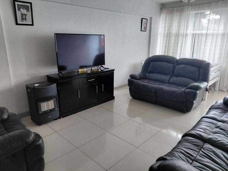A bright living room featuring neutral tiled flooring and large windows with sheer curtains, providing a clean and functional space.