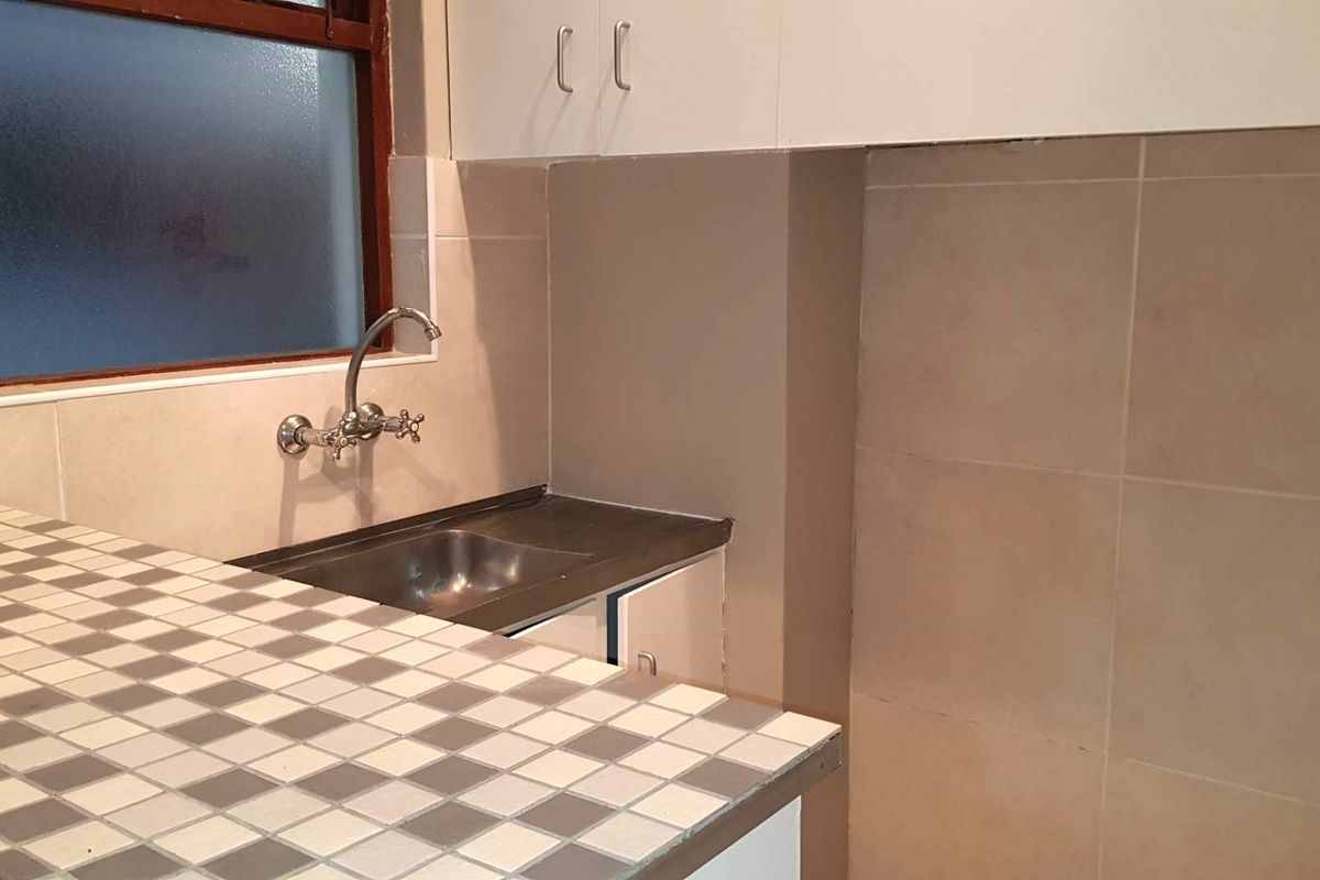 A compact kitchen area featuring a tiled countertop, stainless steel sink, wall-mounted faucet, and built-in cabinetry with a tiled backsplash.