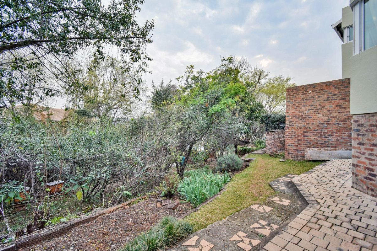 A rear garden area featuring a paved walkway, brick retaining wall, and established garden beds with mature trees and shrubs.