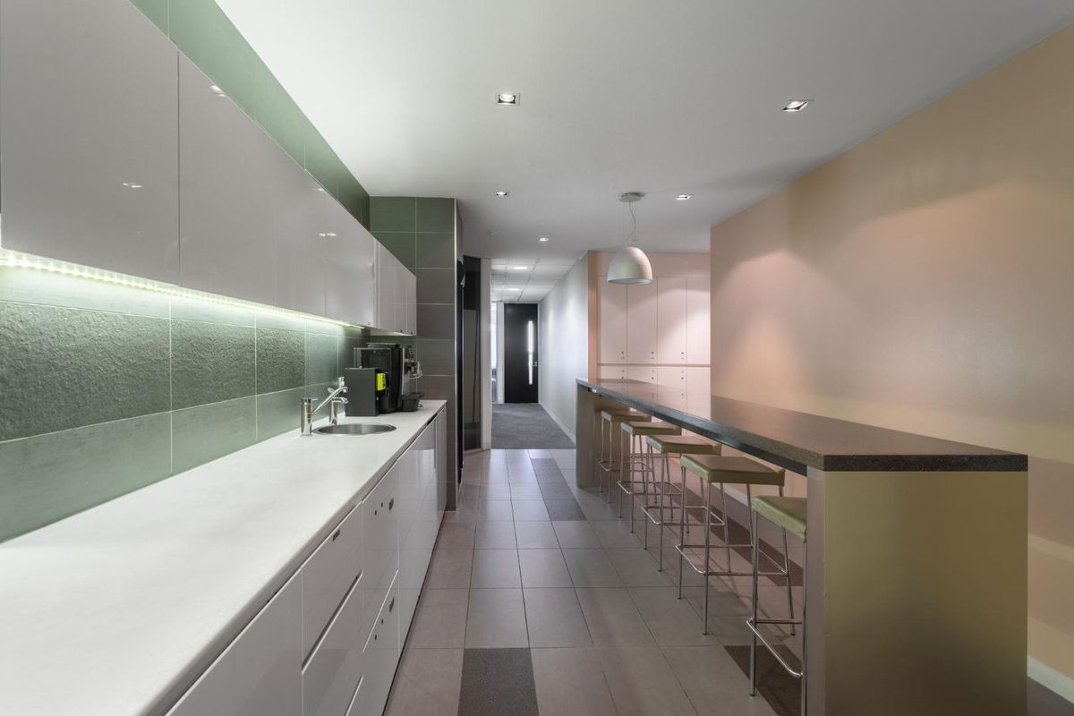 Modern office kitchenette featuring sleek white cabinetry, integrated sink, under-cabinet lighting, and a long breakfast bar with seating.