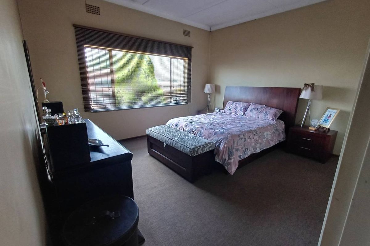 A spacious bedroom featuring neutral wall tones, carpeted flooring, and a large window with fitted blinds providing natural light.