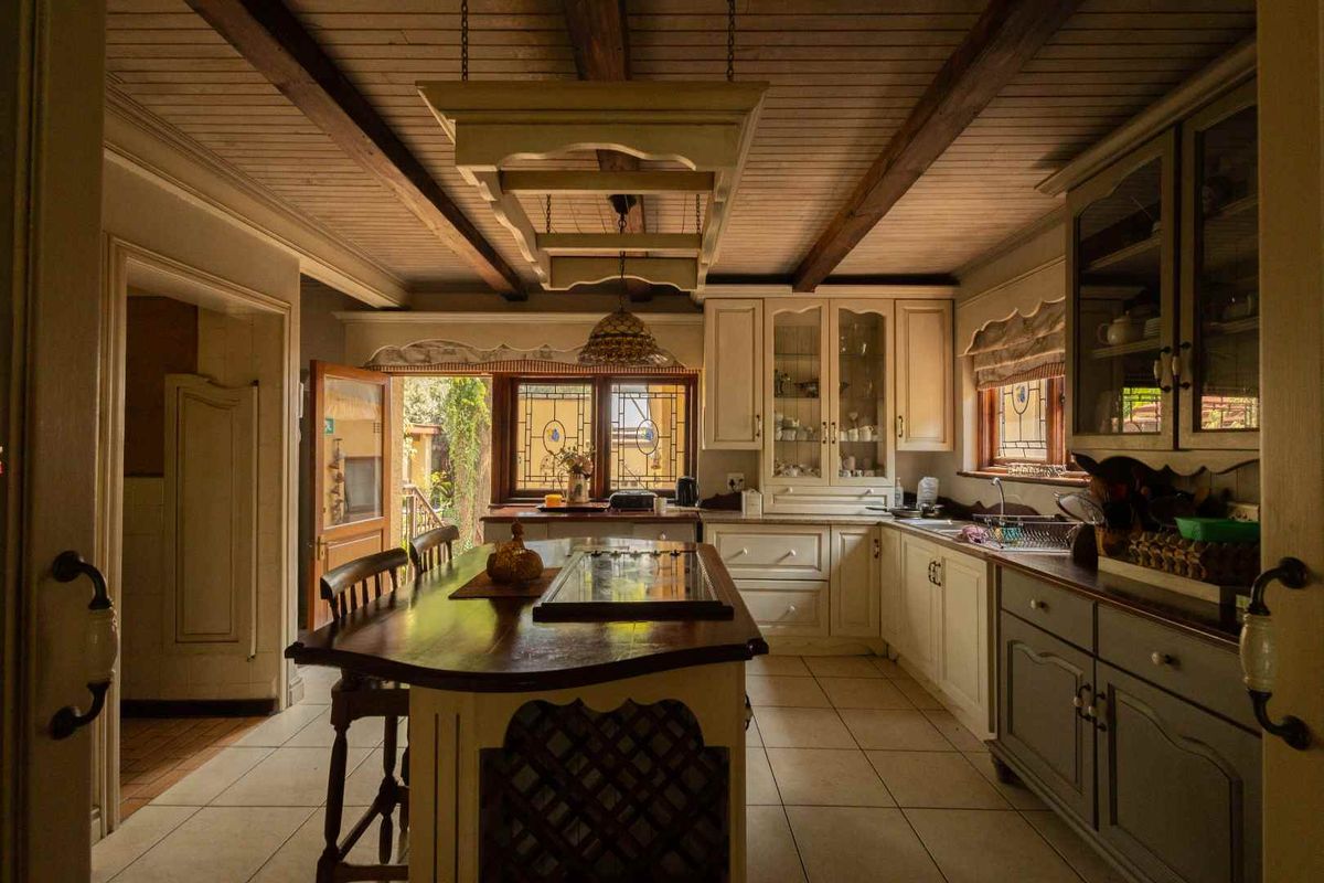 A rustic-style kitchen featuring wooden cabinetry, a central island, exposed ceiling beams, and leaded glass windows overlooking an outdoor area.