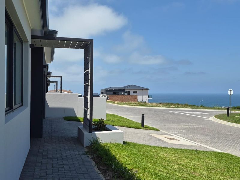 Modern exterior view of the property frontage featuring paved driveway, landscaped lawn, and architectural pergola with scenic coastal views in the background.