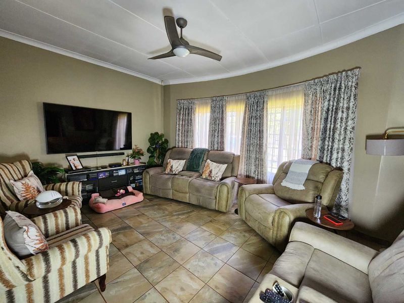 A comfortable living room featuring tiled flooring, a ceiling fan, and large windows with curtain treatments, providing a functional and inviting space.