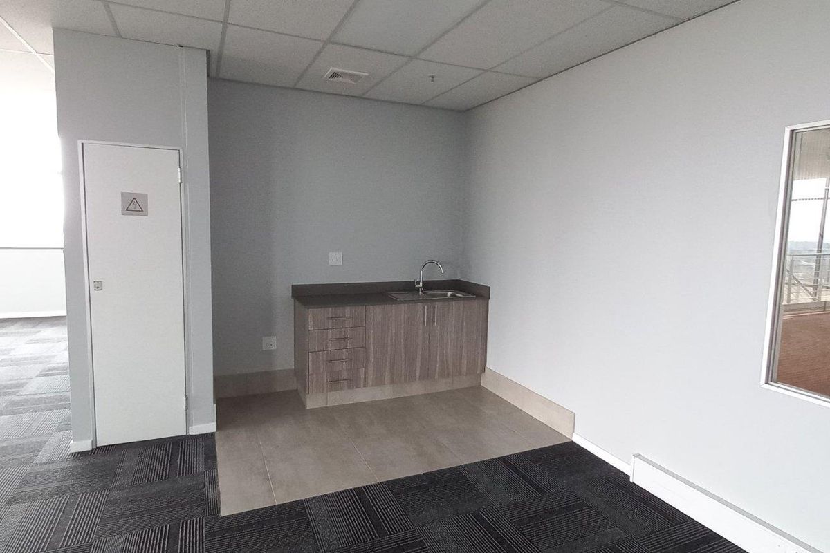 A functional kitchenette area within an industrial office space, featuring built-in cabinetry, a stainless steel sink, and modern carpet tile flooring.