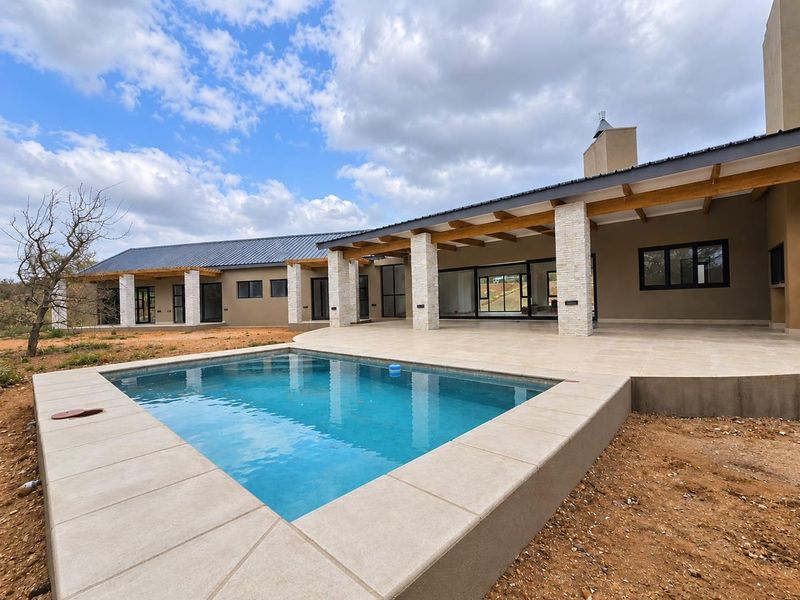 Spacious outdoor pool area featuring a tiled patio, stone-clad pillars, exposed timber beams, and a modern swimming pool set against a natural landscape.