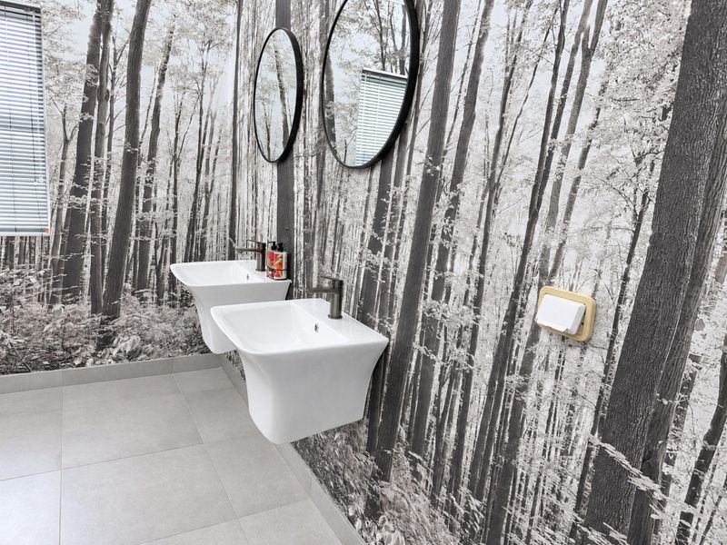 A modern office bathroom featuring wall-mounted double basins, tiled flooring, and a distinctive forest-themed wallpaper finish.