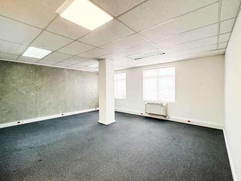 A vacant, open-plan office space featuring a suspended ceiling, recessed lighting, a central structural column, and a wall-mounted HVAC unit.