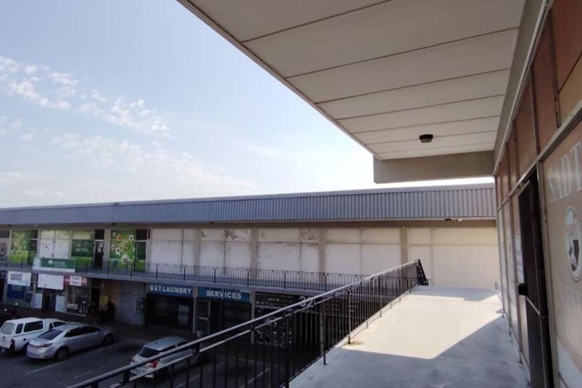 An elevated exterior walkway or balcony area featuring a metal safety railing, concrete flooring, and a protective roof canopy overlooking the parking area.