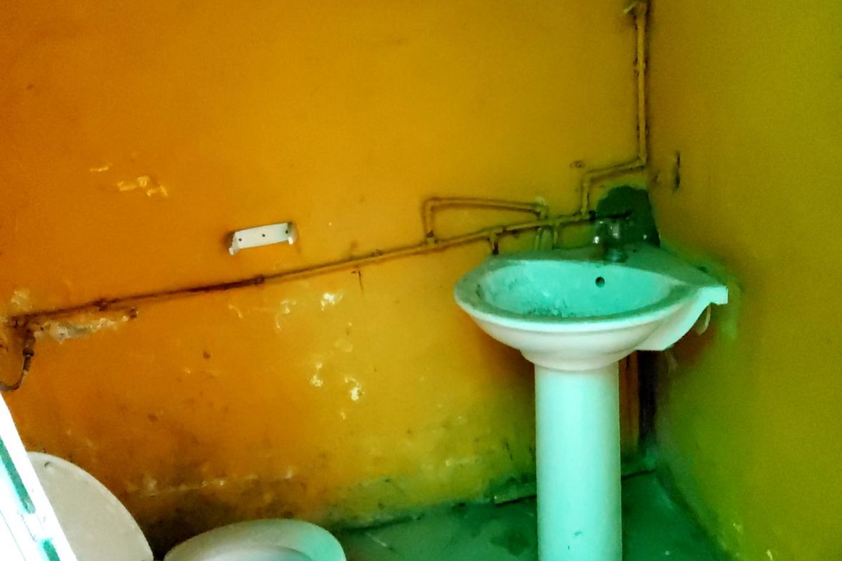 A small, dated bathroom featuring a pedestal sink and toilet with visible wall surface deterioration and exposed plumbing.