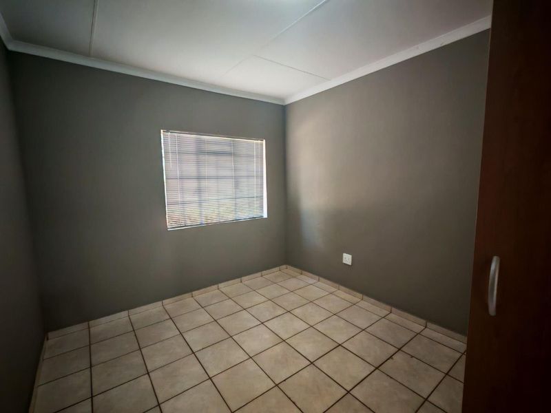 A clean, empty bedroom featuring neutral grey walls, tiled flooring, and a window fitted with venetian blinds.