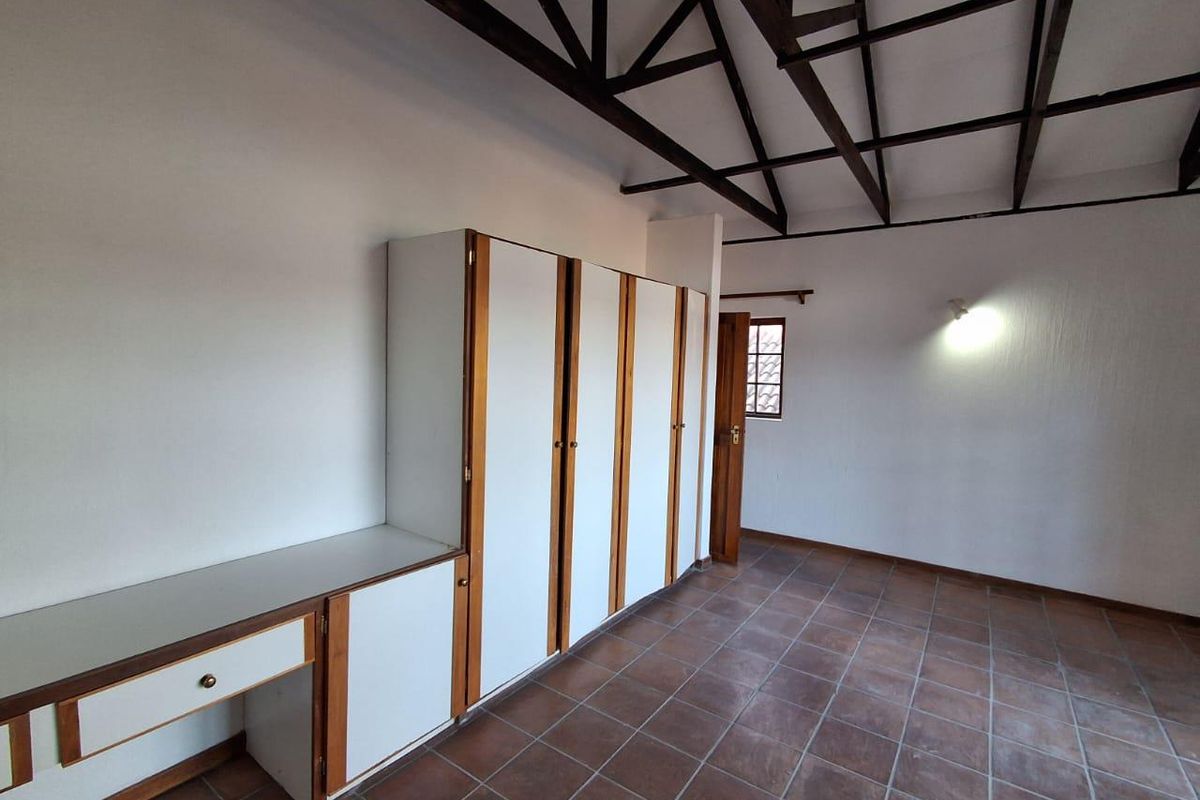 A spacious bedroom featuring built-in cupboards, a built-in desk, exposed timber roof trusses, and tiled flooring.