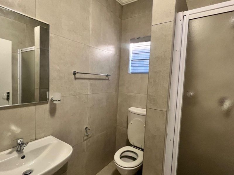 A clean, modern bathroom featuring neutral wall tiles, a pedestal basin, a toilet, and a glass-enclosed shower.