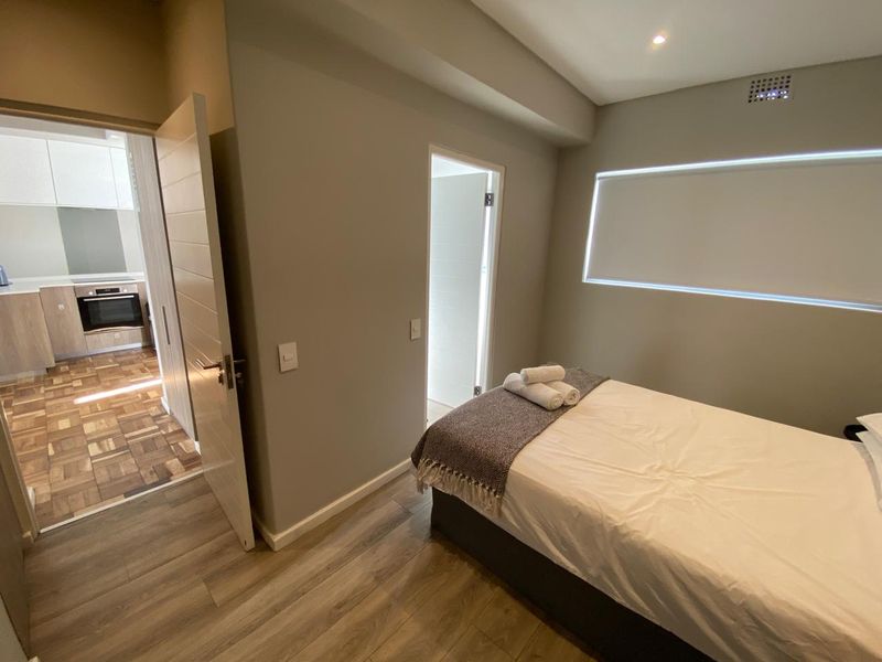 A modern bedroom featuring laminate flooring, recessed lighting, and a roller blind, with direct access to an adjacent kitchen area.