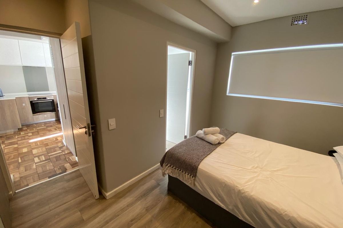 A modern bedroom featuring laminate flooring, recessed lighting, and a roller blind, with direct access to an adjacent kitchen area.