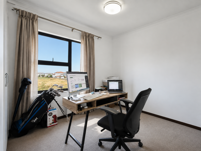 A functional home office space featuring neutral carpet flooring, a large window with curtain rod, and overhead lighting, overlooking an open landscape.