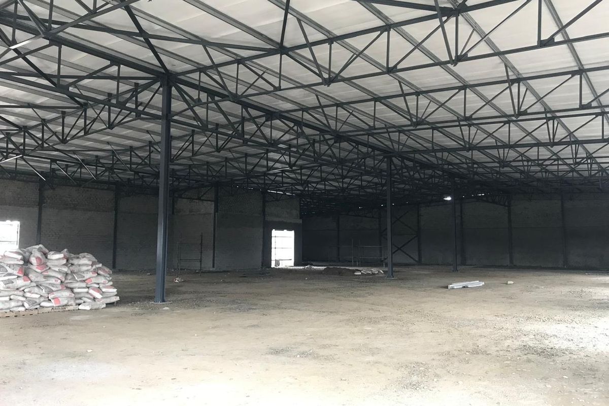 Large open-plan industrial warehouse space featuring exposed steel roof trusses, support columns, and unfinished brick walls with a concrete floor.