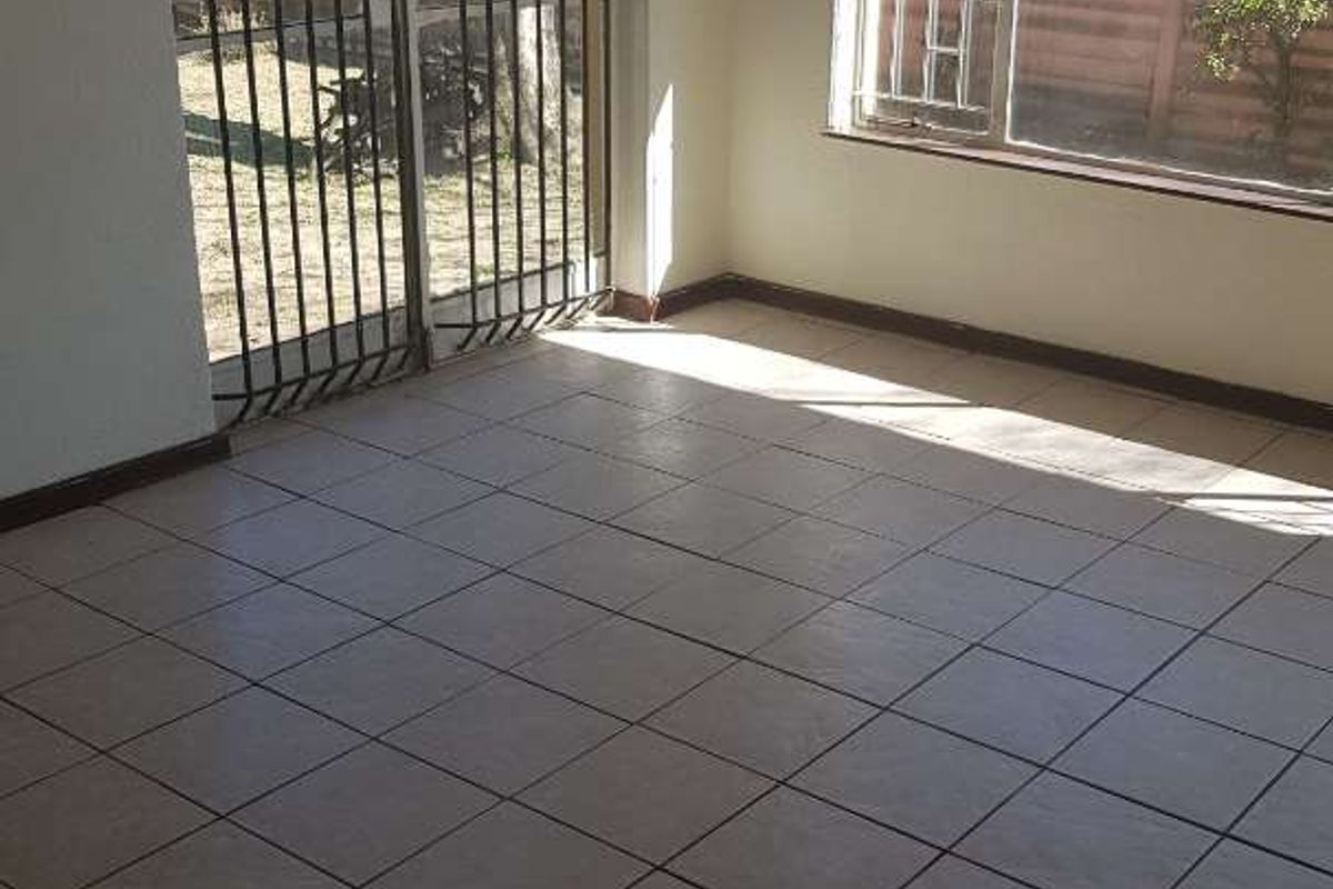 An empty living area featuring neutral floor tiles, large windows with security bars, and a secure metal door leading to the exterior.