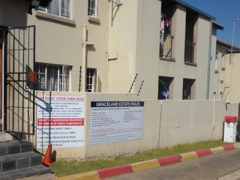 Exterior view of a townhouse complex entrance featuring a security gate, perimeter wall with electric fencing, and paved driveway access.