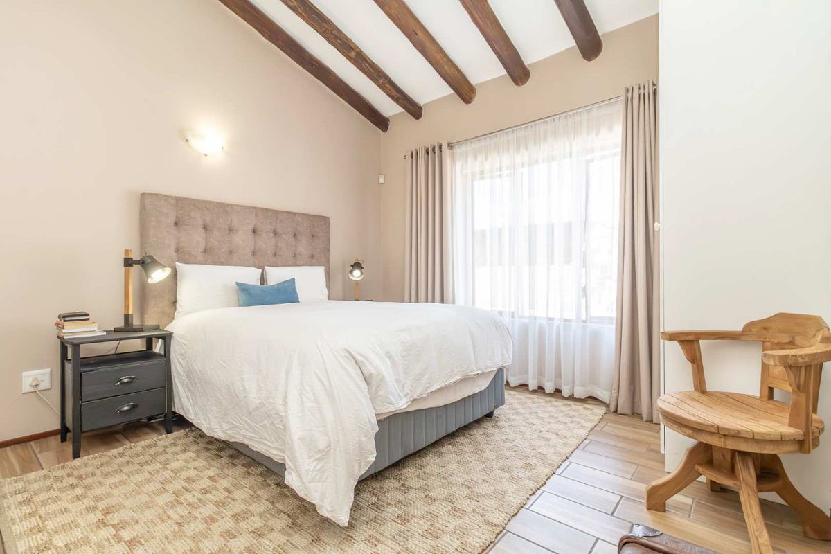 A well-lit bedroom featuring exposed timber ceiling beams, tiled flooring, and large windows with neutral curtains.