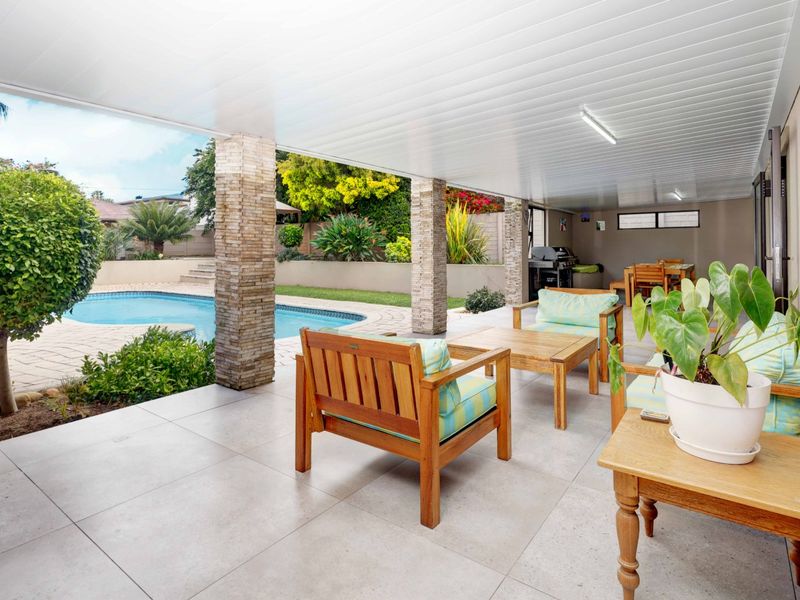 A spacious covered patio featuring stone-clad columns and tiled flooring, overlooking a private swimming pool and landscaped garden area.