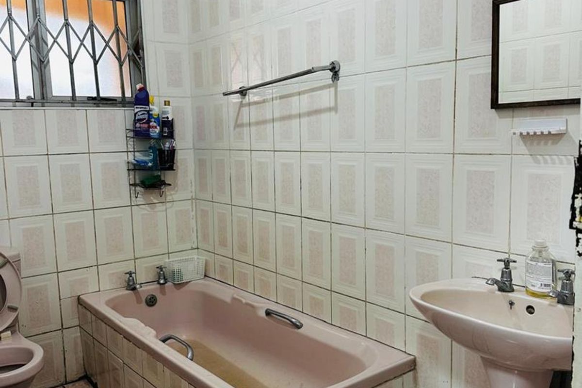 A dated bathroom featuring a pink bathtub and pedestal basin, tiled walls, and a security-grilled window.