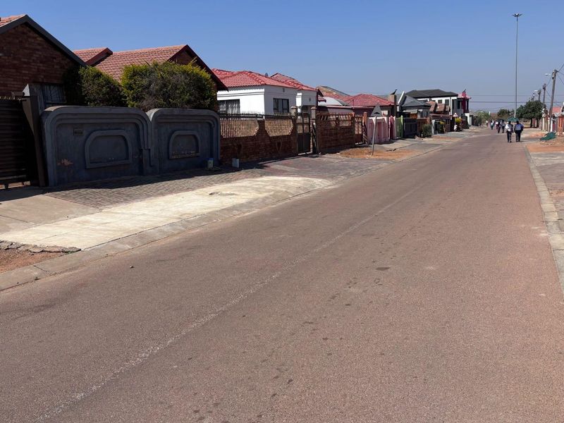 A street view showing the residential neighborhood approach, featuring paved roads, sidewalks, and boundary walls of the surrounding properties.