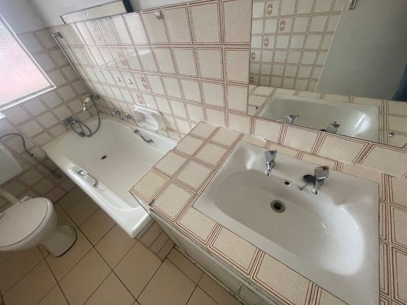A dated bathroom featuring a tiled vanity with inset basin, a built-in bathtub with shower mixer, and a toilet.