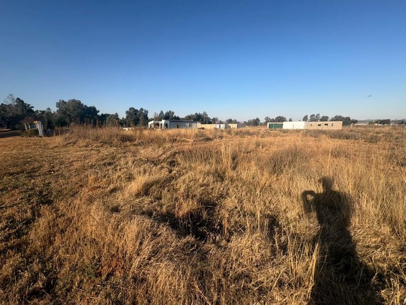 A wide-angle view of a large, undeveloped residential plot featuring dry, overgrown grass and distant existing structures.