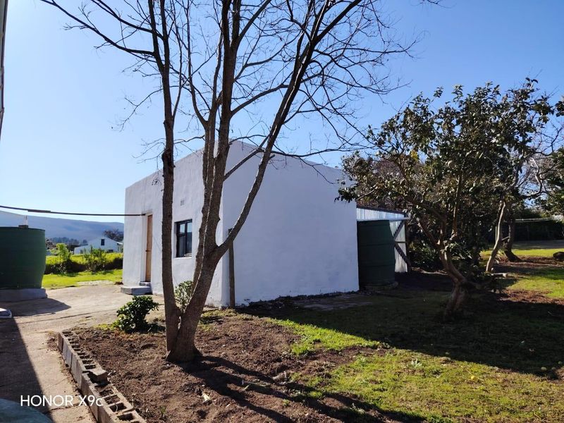 Rear view of a detached outbuilding featuring white rendered walls, a metal roof, and an adjacent water tank set within a garden area.