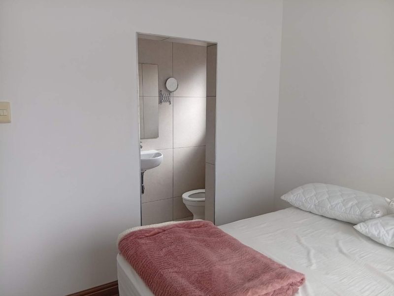 A bedroom featuring an open-plan en-suite bathroom with a wall-mounted basin and toilet, finished with neutral wall tiles.