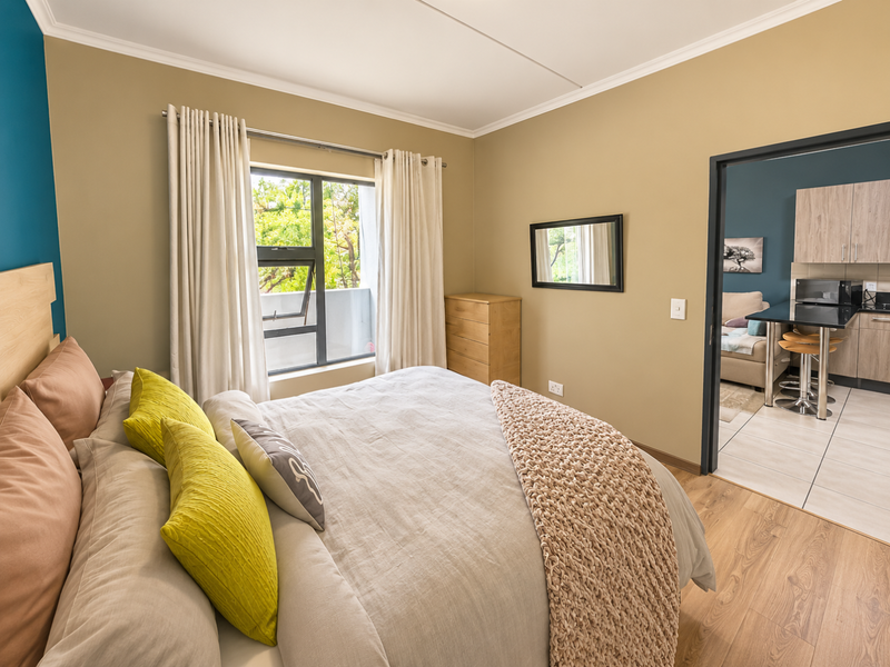 A well-maintained bedroom featuring laminate flooring, neutral wall tones, and an adjoining view into the kitchen area.