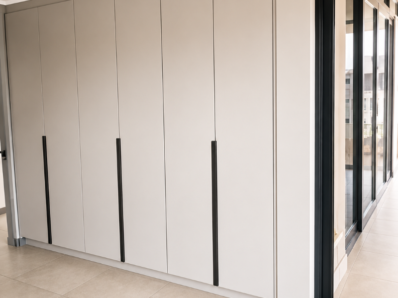 A modern bedroom interior featuring sleek, floor-to-ceiling built-in cupboards, neutral tiled flooring, and a large sliding glass door leading to an outdoor area.