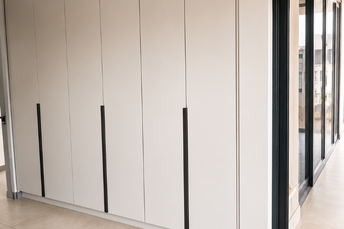 A modern bedroom interior featuring sleek, floor-to-ceiling built-in cupboards, neutral tiled flooring, and a large sliding glass door leading to an outdoor area.