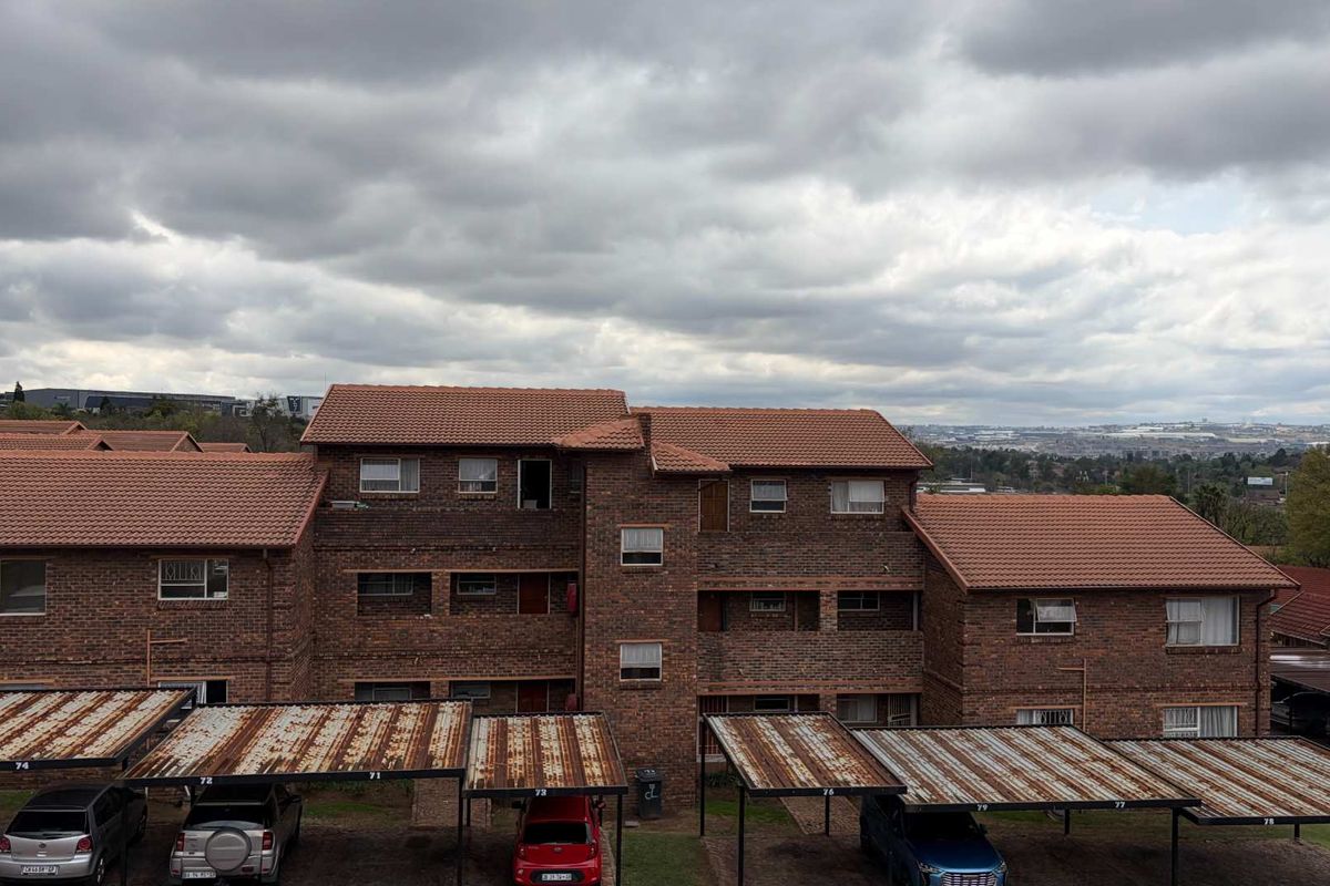 Exterior view of a multi-story face-brick townhouse complex featuring tiled roofing and multiple carport parking bays with visible surface corrosion.