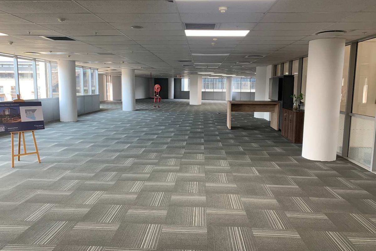 Spacious open-plan office area featuring carpet tiles, structural columns, large windows for natural light, and a suspended ceiling with recessed lighting.