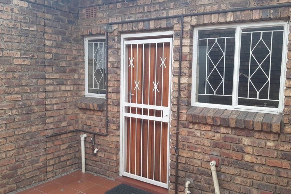 A rear exterior view of the townhouse featuring face brick walls, a security gate, and tiled patio area with visible external plumbing.