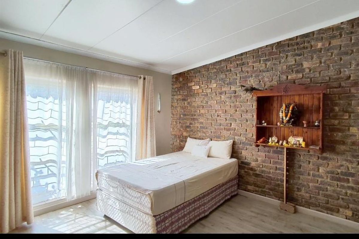 A bright bedroom featuring a distinctive exposed brick feature wall, light-toned flooring, and large windows allowing for ample natural light.