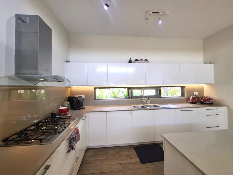 Modern kitchen featuring gloss white cabinetry, stone countertops, gas hob, stainless steel extractor, and a unique horizontal window splashback overlooking the garden.