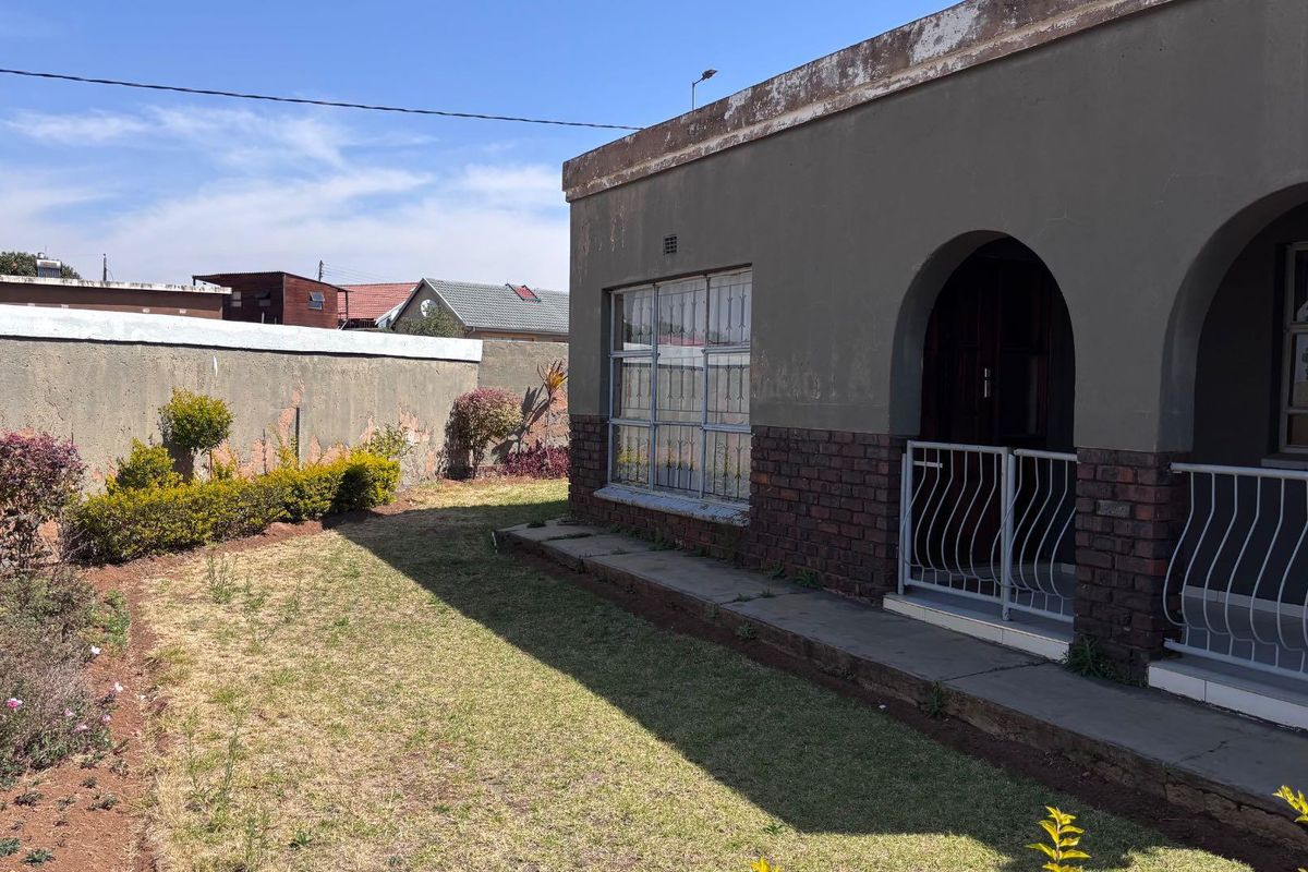 Rear exterior view of the house featuring a brick and plaster facade, arched entrance, security bars, and a private garden area with perimeter wall.