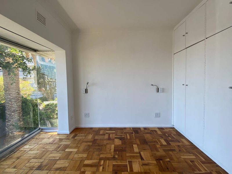 A bright, empty bedroom featuring classic parquet flooring, ample built-in cupboard storage, and wall-mounted reading lights, with a large window overlooking greenery.