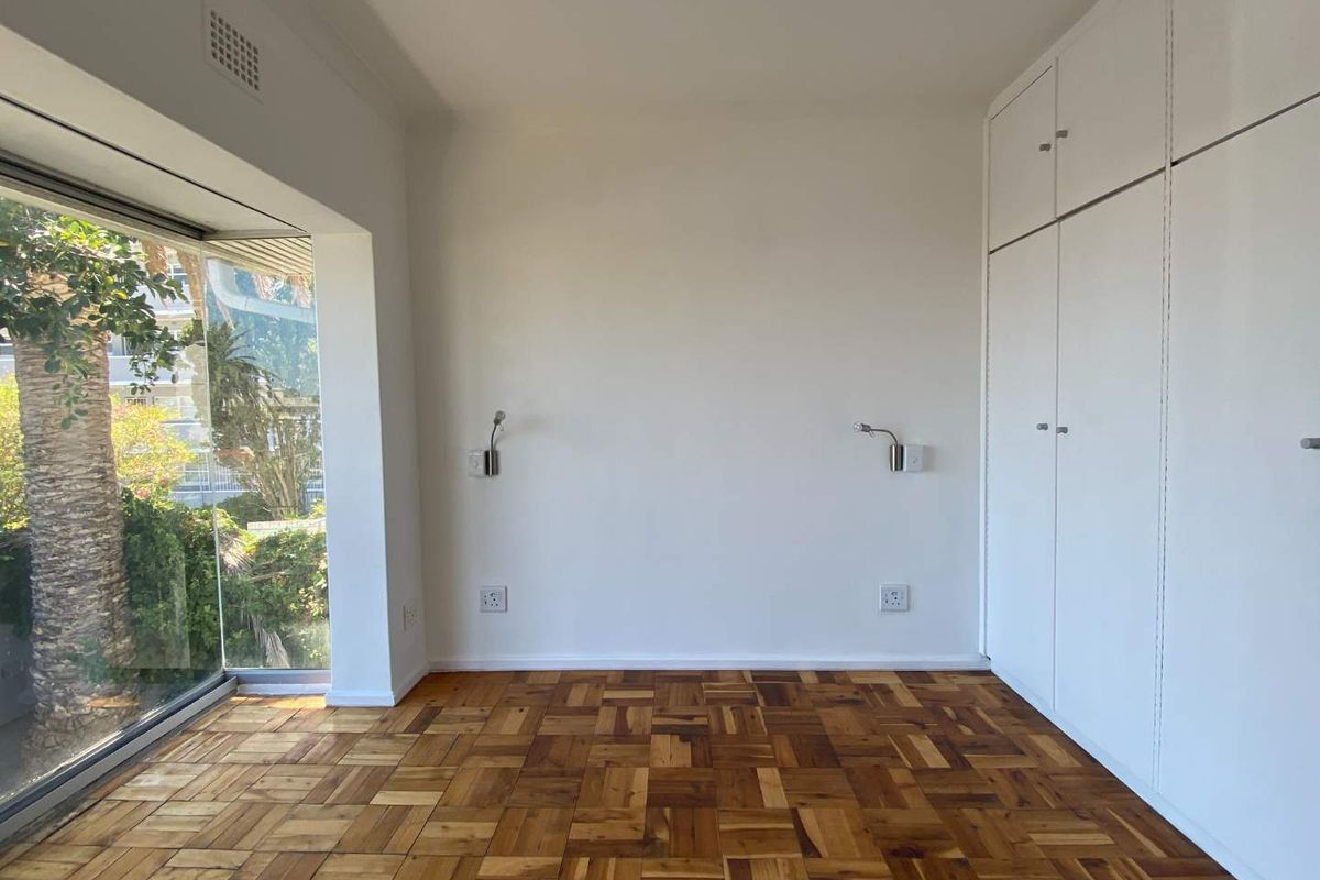 A bright, empty bedroom featuring classic parquet flooring, ample built-in cupboard storage, and wall-mounted reading lights, with a large window overlooking greenery.