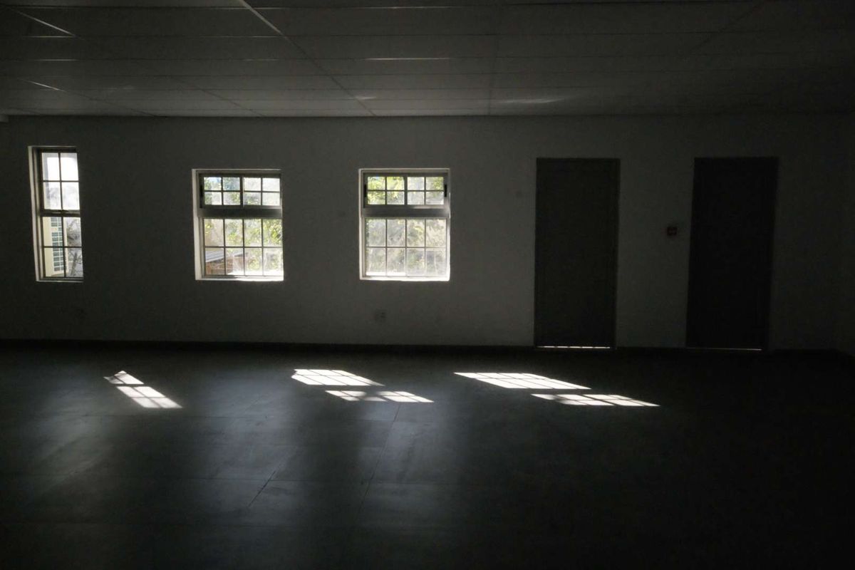 A spacious, empty office area featuring tiled flooring, a grid ceiling, and multiple windows providing natural light.