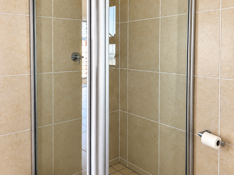A clean, functional corner shower unit with glass enclosure, tiled walls, and floor, featuring a modern shower mixer and wall-mounted toilet paper holder.