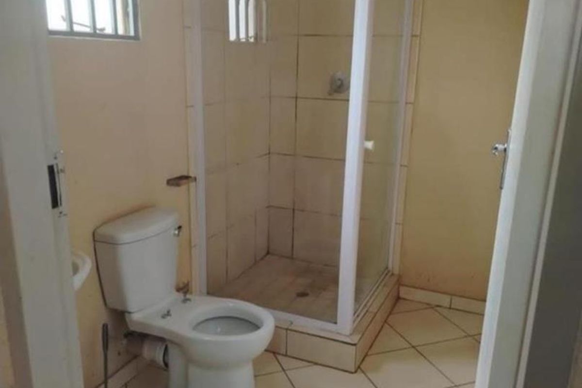 A functional bathroom featuring a corner shower enclosure, a standard toilet, and tiled flooring with security bars on the windows.